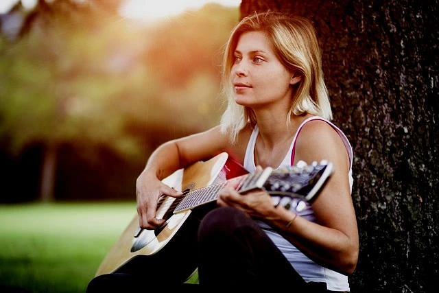 Guitarist leaning on tree in the park as they play.